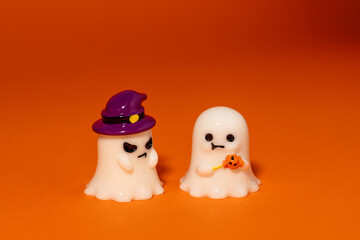One ghost wears a purple witch hat and looks angry on second ghost, while he is holding pumpkin candy a looks calm on vibrant orange background