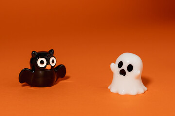 Playful Halloween scene featuring a small ghost figurine scaring a black owl toy against an orange background.