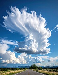 A long road stretches to a puffy, large cloud formation in a bright blue sky. Desert landscape fades into distance