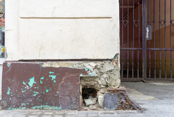Damaged building facade with peeling plaster, visible foundation cavities, exposed bricks, corrosion, metal entrance gate visible in the background.
