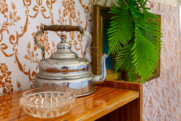 PRL interior with aluminum kettle, glass bowl, and green fern placed on wooden shelf against patterned wallpaper background.