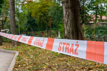 Warning tape with Polish inscription “STRAŻ” securing area near trees and sidewalk, marking restricted outdoor emergency scene.
