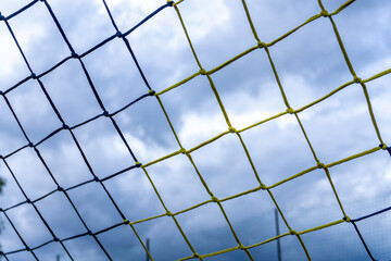 Football goal net made of yellow and black ropes against cloudy sky background, photographed from sports field perspective.
