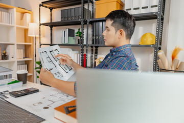 Young male architect working thoughtfully at office desk, analyzing house blueprints and...