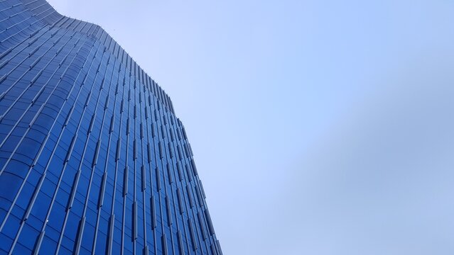Upward view of a sleek, reflective glass skyscraper with geometric patterns, rising against a bright, open blue sky in an urban setting.