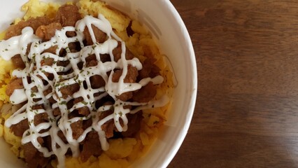 Top view of a rice bowl topped with seasoned meat and mayonnaise sauce, served in a white container over a wooden table.  
