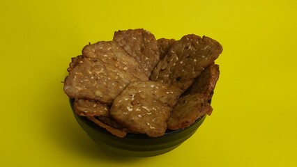 Fried tempeh chips in bowl on yellow background  
