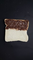 A slice of white bread partly covered with chocolate spread placed diagonally on a black surface with small scattered crumbs.  
