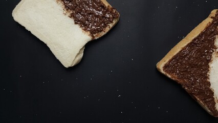 Two slices of bread partly covered with chocolate spread, placed diagonally on a black surface with scattered crumbs.  
