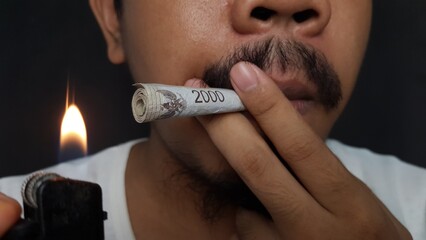 Close-up of a man lighting a rolled rupiah banknote like a cigarette, symbolizing wasting money,...