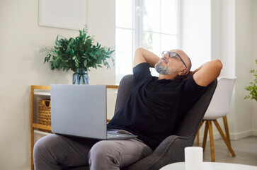 Happy senior man leaning back in comfy armchair to relax and rest at laptop during coffee break. Older lazy person in glasses sitting in chair to pause online work in casual living room at home