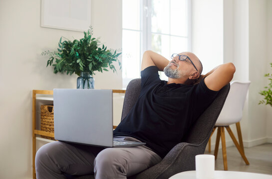 Happy senior man leaning back in comfy armchair to relax and rest at laptop during coffee break. Older lazy person in glasses sitting in chair to pause online work in casual living room at home