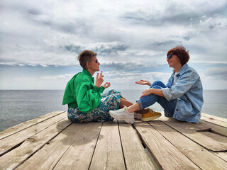Two friends enjoying a deep conversation on a wooden pier by the sea under a cloudy sky