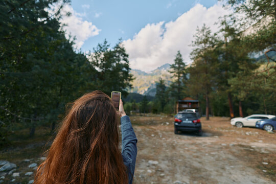 A person with long hair stands in a forest clearing, holding a smartphone to photograph a distant car on a dirt road, as tall trees frame the sunny outdoor scene. - Powered by Adobe