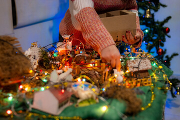 Person in a knitted sweater arranging a Christmas nativity scene, with colorful lights and a Christmas tree in the background.