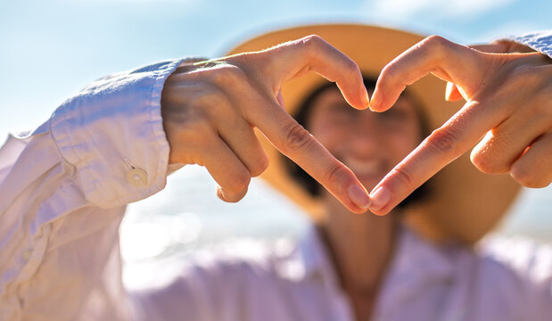 A girl made a beautiful heart with her finger. A cheerful girl makes a heart shape with her hands against a backdrop of sea and sky. - Powered by Adobe