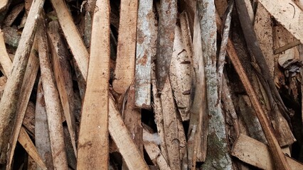 A close-up view of a pile of wooden planks, bark, and rough cut timber stacked together. The wood pieces vary in texture, size, and color, with natural weathering and dirt visible on the surface.  
