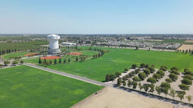 Tracking aerial shot passing water tower, green fields, trees, and residential area.