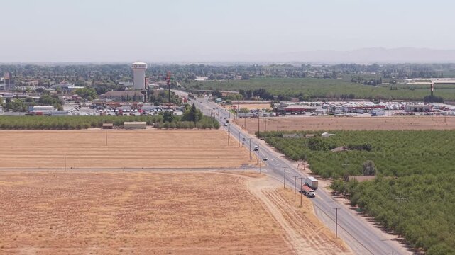Aerial reverse shot following road between fields, leading toward city and water tower in Ripon California.