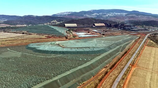 Aerial view of a large quarry mine in Brazil, showing tailings, processing facilities, and surrounding nature with mountains in the distance under clear skies