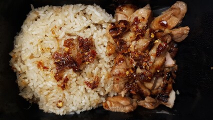 A serving of grilled chicken slices paired with steamed rice and sprinkled chili flakes, presented in a simple black container.    © Ahmad