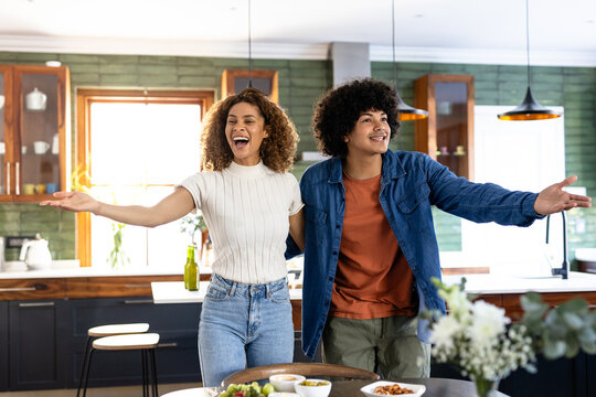 Young couple joyfully welcoming guests in modern kitchen, arms open wide