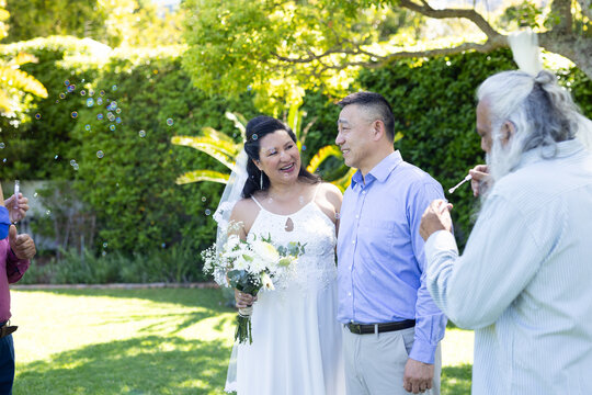 At outdoor celebration, senior couple smiling with friends blowing bubbles in garden