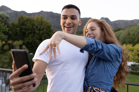 Young couple taking selfie outdoors, showing engagement ring with joyful expressions