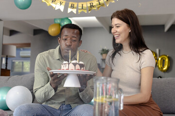 Man blowing out candles on birthday cake while woman smiling beside him