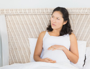 Pregnant Asian woman resting on bed, thoughtfully holding her belly at home