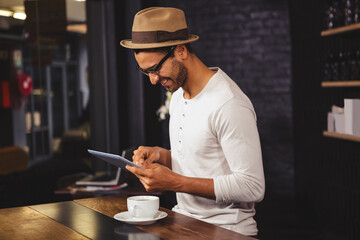 Smiling man in hat using tablet at coffee shop, enjoying his drink