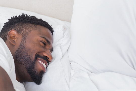 African American man smiling while relaxing comfortably on white bed linens, copy space