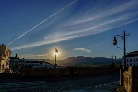 Sun rising behind mountains, illuminating street lamps on cobblestone bridge at dawn in ronda, spain