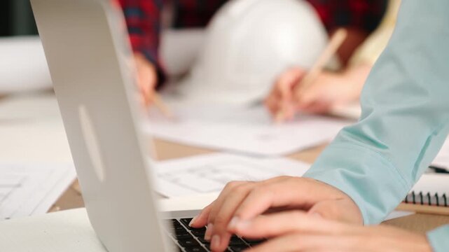 Architect and safety engineer discussion about building blueprint displaying on laptop on table with architectural document and wooden block scatter around at modern office. Closeup. Delineation.