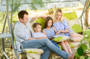 Close-up of family sitting together on garden swing, enjoying time together in backyard. Mom, dad, girl, and boy are reading book together, discussing story they've read. Vacation out of town