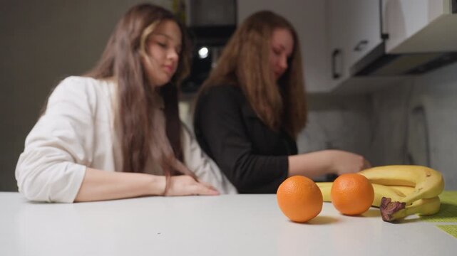 Friends in kitchen pack fruits from table, chatting and smiling while placing oranges and bananas into bag, warm daylight, soft blur background, relaxed preparation mood, casual moment before picnic