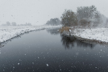 A peaceful winter scene with snowflakes falling on a calm river, surrounded by snowy trees and fields under a gray sky.