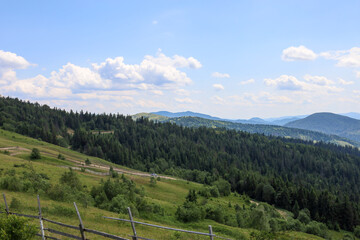 Fototapeta premium ​A serene summer day in the mountains, where green slopes are covered with dense forests and winding trails. Fluffy white clouds drift lazily across the endless blue sky, adding to the grandeur of thi