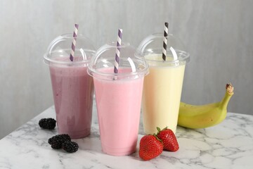 Milkshakes of different flavors in plastic cups and ingredients on white marble table, closeup