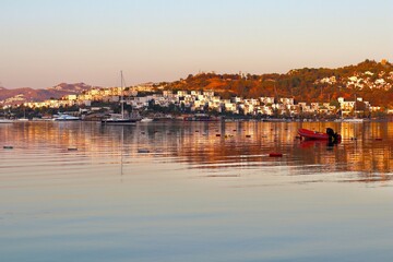 Fototapeta premium A stunning sunrise over a calm, reflective bay, with sailboats moored in the glassy water. The sun's first rays illuminate the mountains and the whitewashed buildings of a coastal town on the hillside