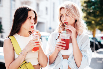 Two young beautiful smiling glam female in trendy summer clothes. Sexy women posing outdoors. Positive models holding and drinking fresh cocktail smoothie drink in plastic cup. Bright makeup, red lips