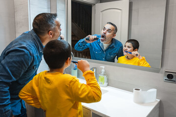 Father teaching oral hygiene to his son, brushing teeth together in bathroom, making funny faces in the mirror