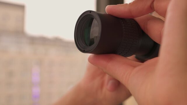A man is intently observing the world outside his window with binoculars, showing curiosity