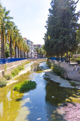 Fototapeta premium Urban river with dirty water, algae, and scattered litter in a modern city. Environmental pollution problem caused by urbanization and lack of waste management. Concept of climate change, water