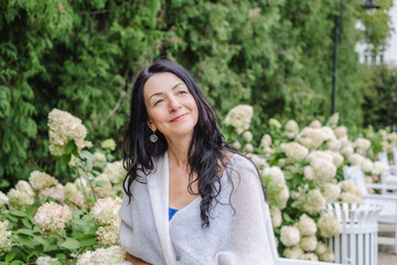 Elegant elder woman with long dark hair, wrapped in a soft shawl, enjoys a serene moment in a beautiful garden surrounded by blooming flowers, embodying wisdom and natural beauty