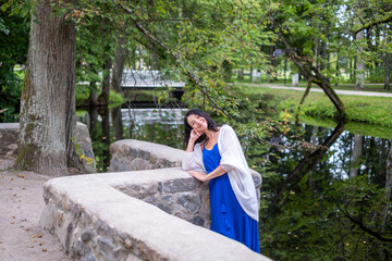 Fototapeta premium Woman in blue dress leans against stone wall by serene water, surrounded by lush greenery, embodying tranquility, beauty, and graceful femininity in a peaceful natural setting
