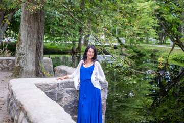 Woman in blue dress stands gracefully by serene water, surrounded by lush greenery, embodying...