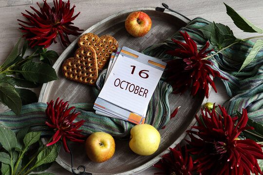 Calendar for October 16: the number 16 and the month's name "October" in English on a flip calendar among dahlias, cookies, and fruit