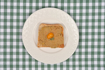 A top-down shot of a piece of toast with homemade chicken liver pate, garnished with a small yellow tomato, on a plate with a green gingham tablecloth