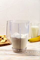 Banana milkshake. Blender containing creamy smoothie ingredients is placed on wooden table, accompanied by fresh bananas and glass of smoothie, creating inviting kitchen scene. Banana smoothie